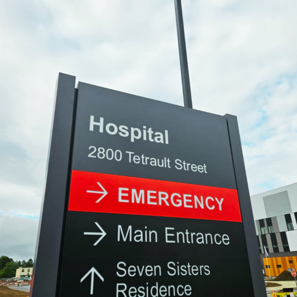 Tall pylon sign located at the entrance of a hospital, displaying the facility's name and directional information for emergency, and main entrance.